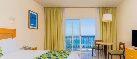 A hotel room with a bed, a desk with a chair, a TV, and a balcony with a view of the sea.