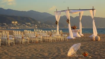 Ein Strandhochzeitsaufbau mit Reihen weißer Stühle, einem Baldachin und einer Kulisse aus Bergen und Meer.