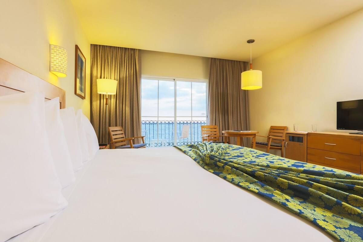 Habitación de hotel con cama, escritorio, silla, televisión y vista al mar.