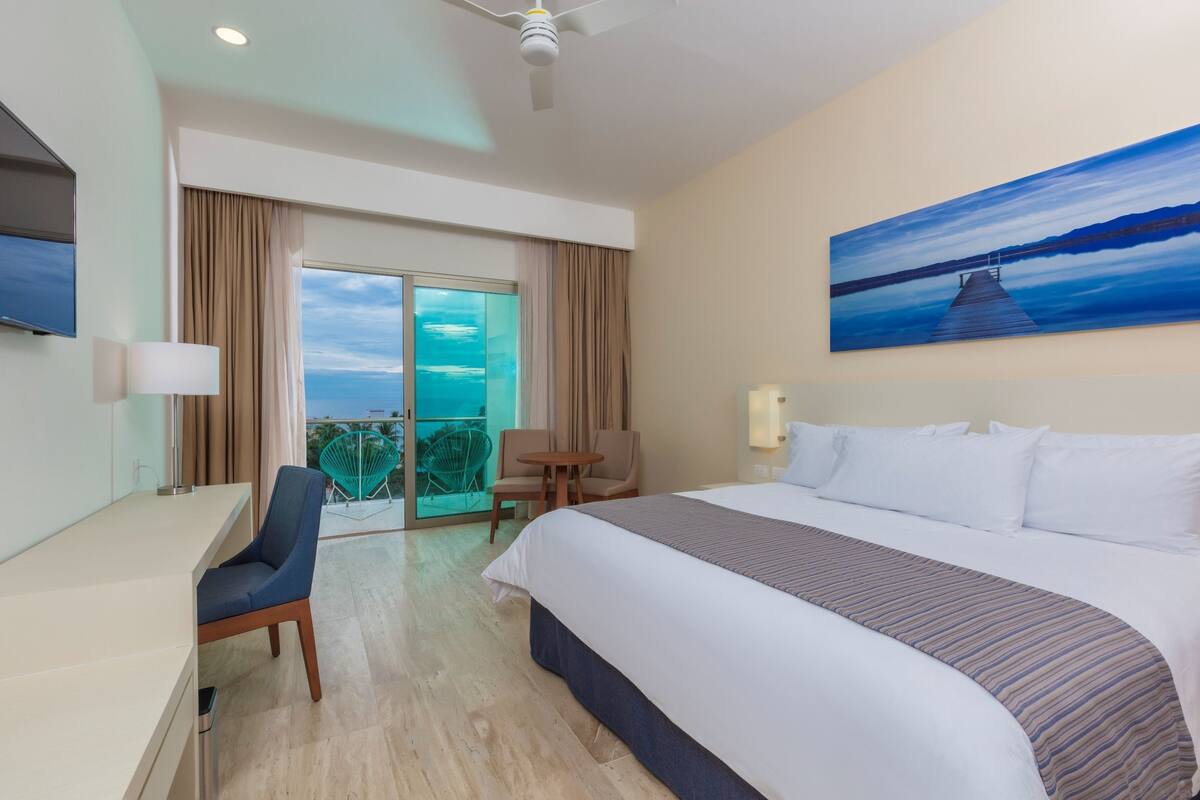 Habitación de hotel con cama, escritorio, silla, balcón con vistas y un cuadro en la pared.
