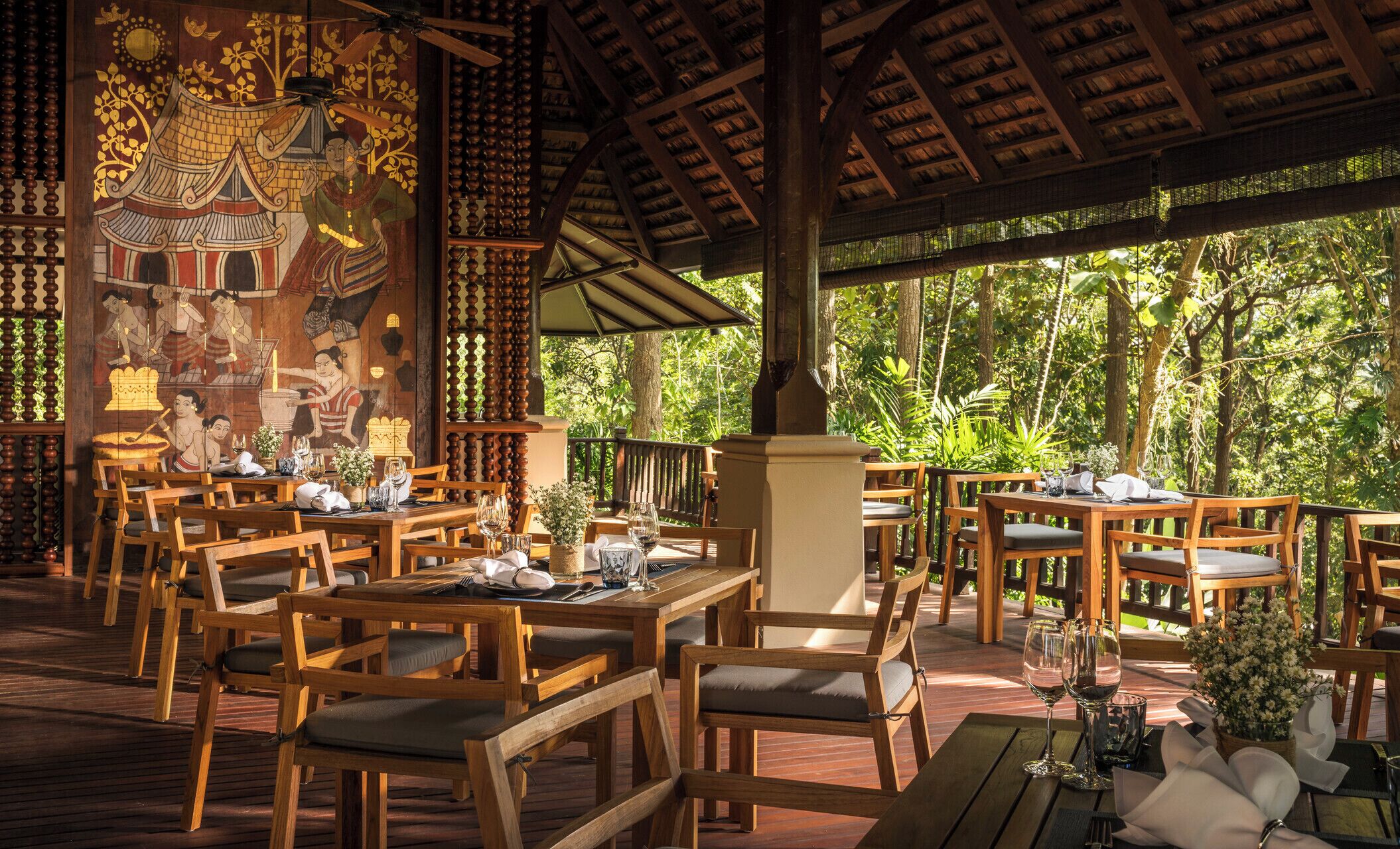 An outdoor restaurant with wooden tables and chairs, surrounded by lush greenery.
