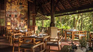 An outdoor restaurant with wooden tables and chairs, surrounded by lush greenery.