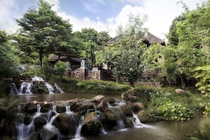 A waterfall cascading over rocks into a pond, surrounded by lush greenery and traditional buildings.