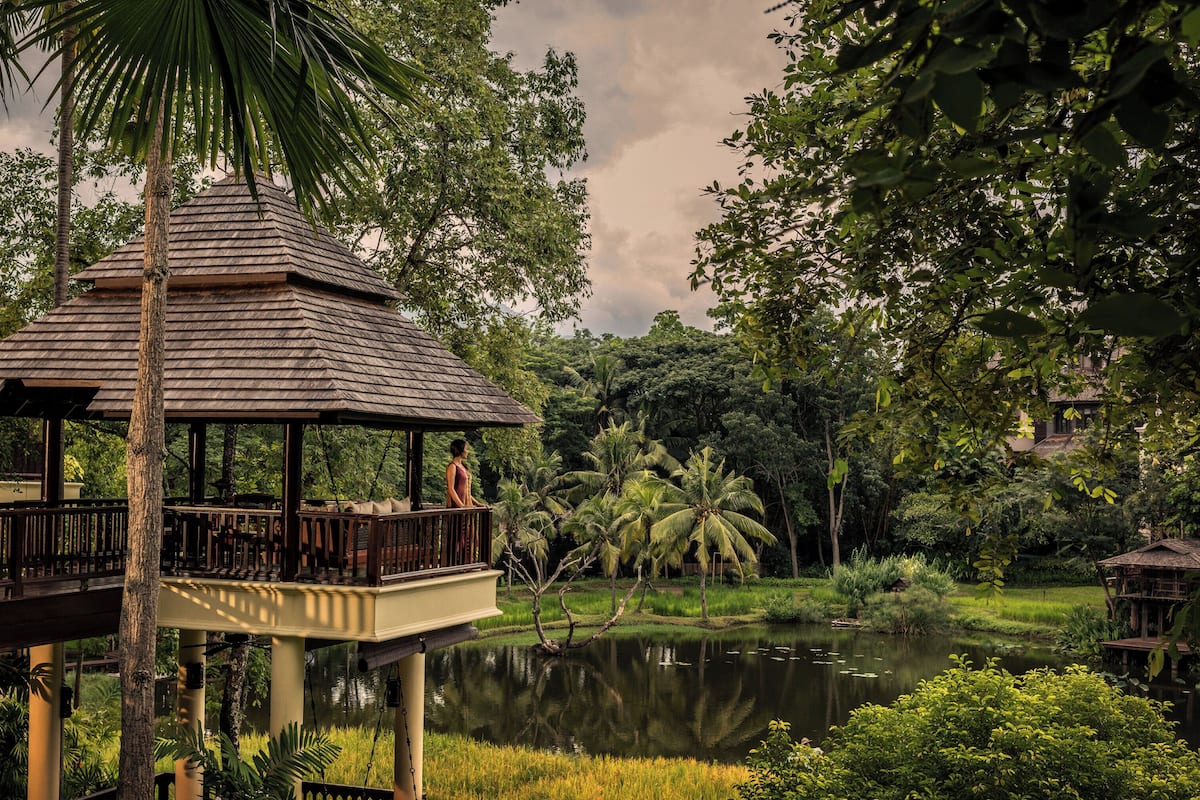 A wooden gazebo with a balcony overlooking a pond, surrounded by lush greenery and palm trees.