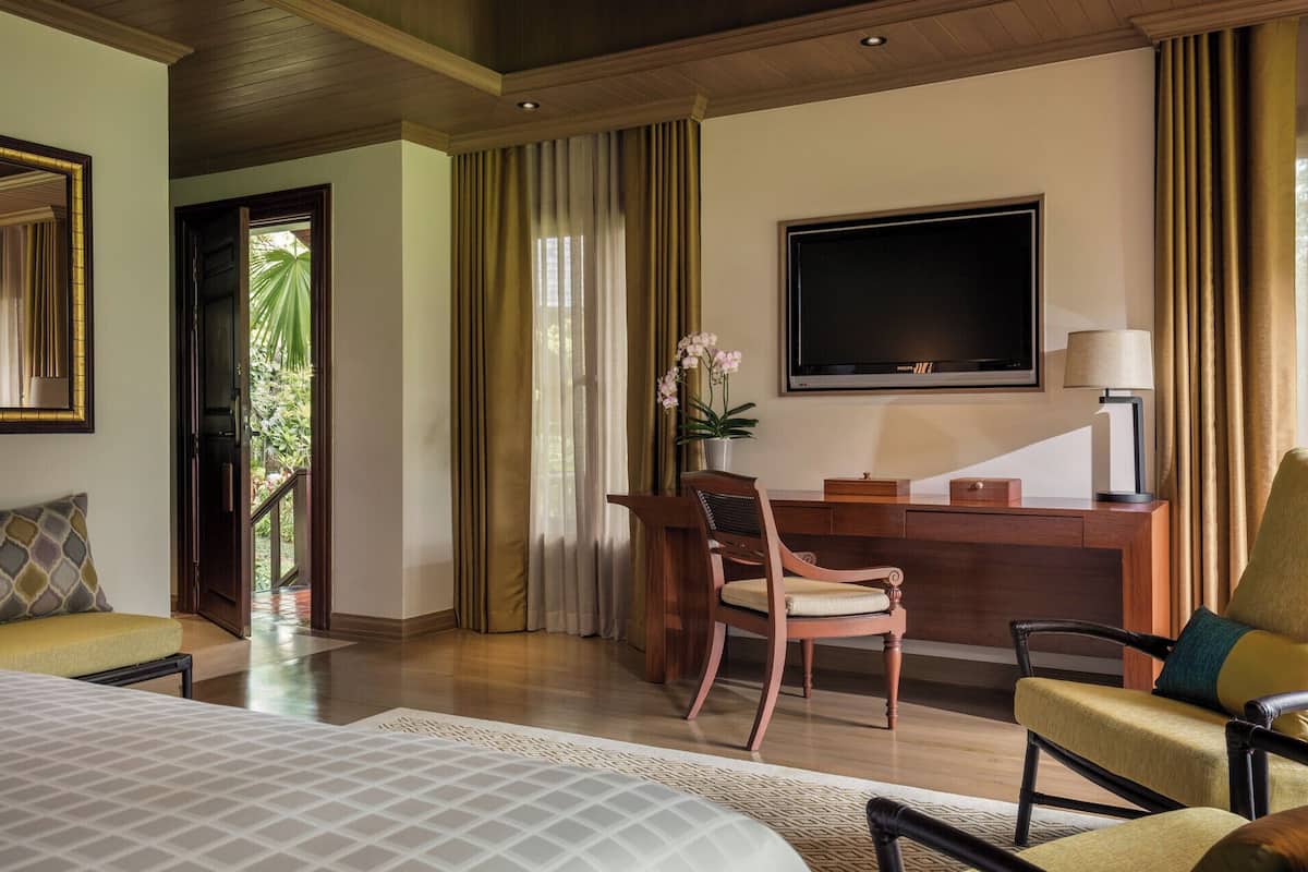 A hotel room with a bed, two chairs, a desk with a lamp, a TV, and a view of greenery outside.