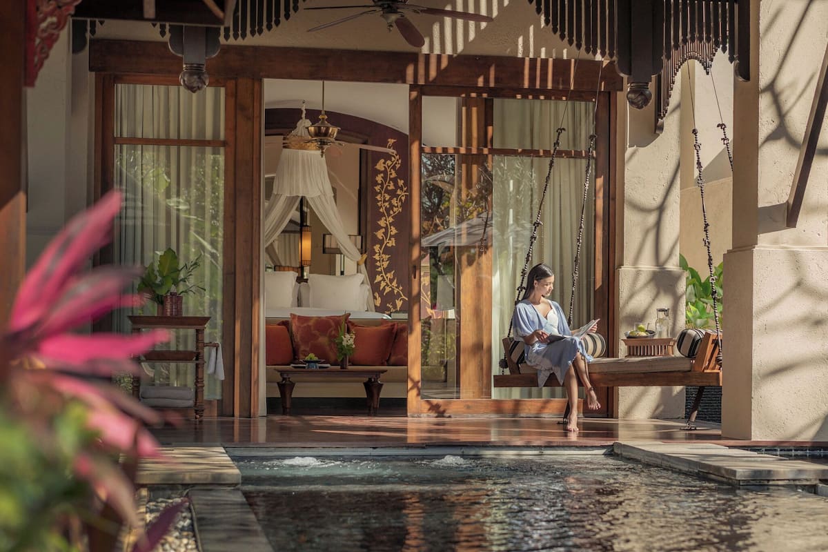 A serene indoor pool area with a person relaxing on a swing chair, surrounded by wooden furniture and a decorative hanging lamp.