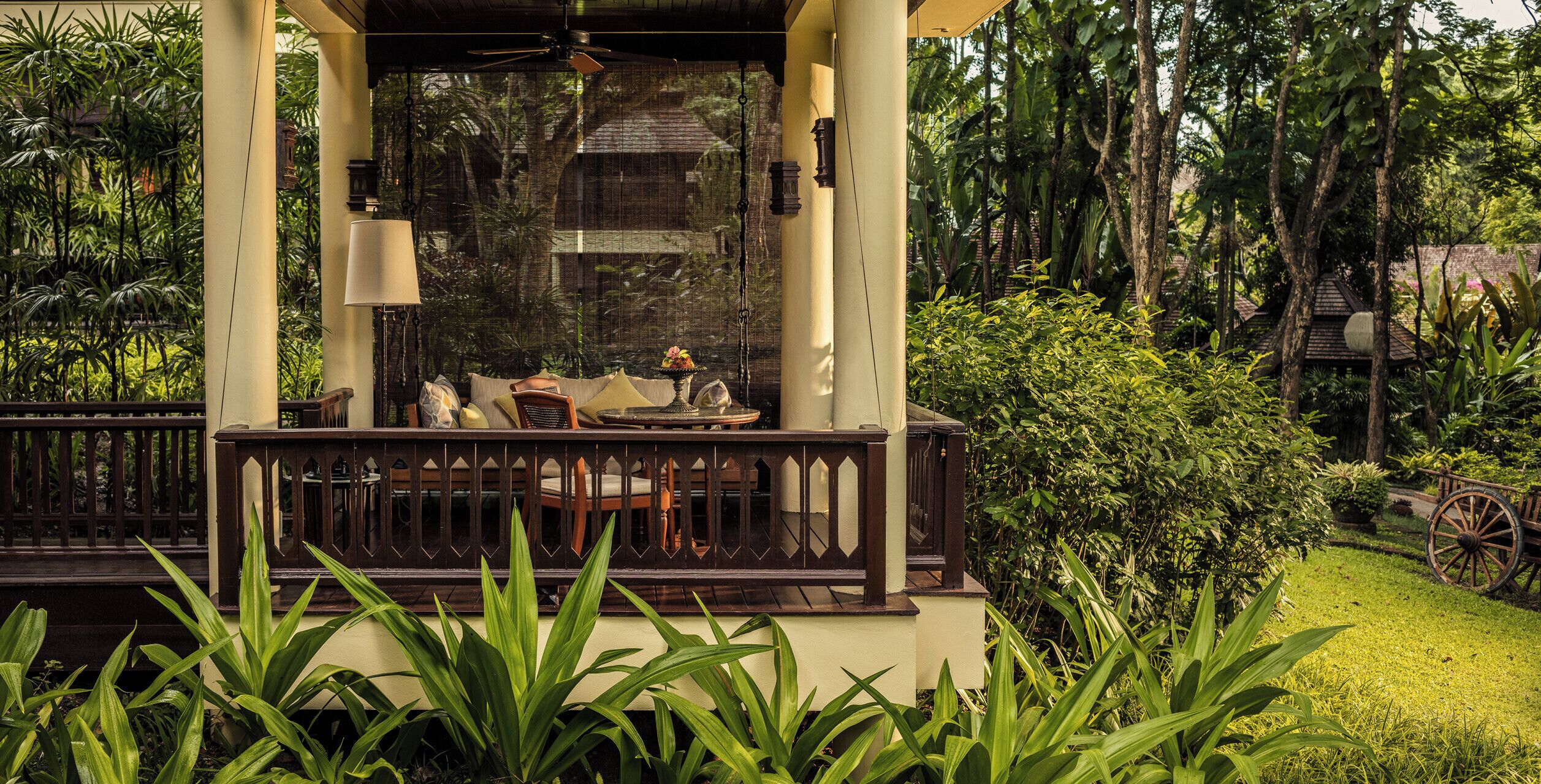 A covered outdoor seating area with a table and chairs, surrounded by lush greenery and a traditional wooden cart.