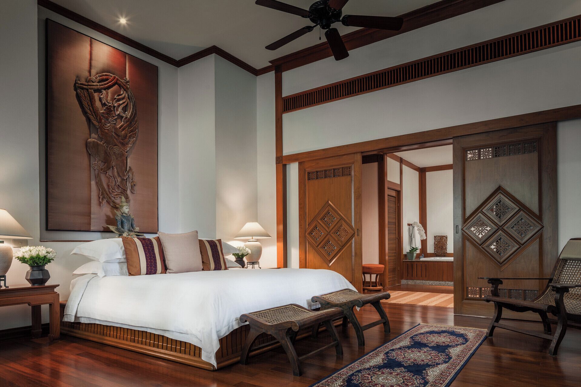 A spacious bedroom with a large bed, wooden flooring, a ceiling fan, and a decorative wall piece.