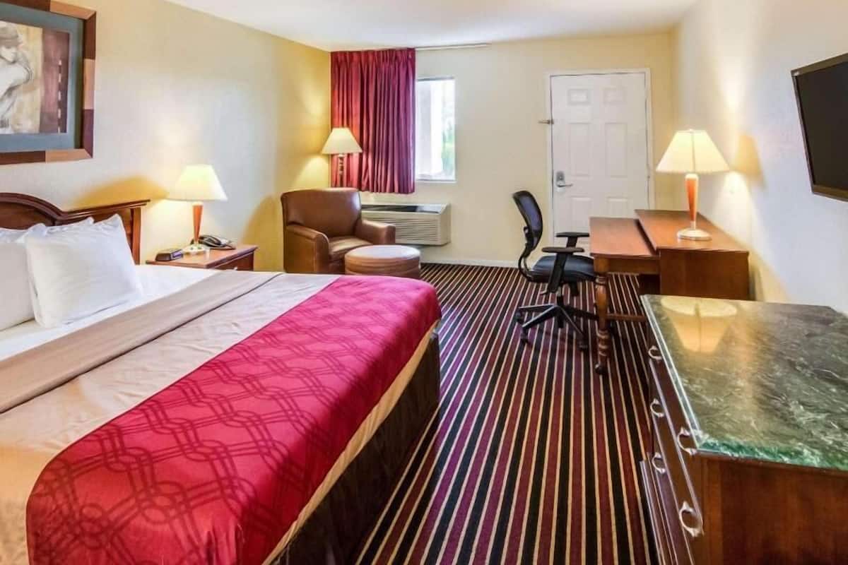 A hotel room with a bed, a desk, a chair, a lamp, and a painting on the wall.
