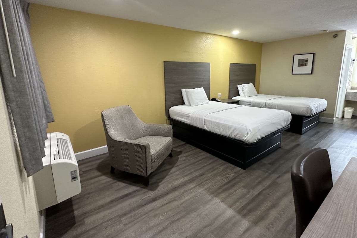 A hotel room with two beds, a chair, a desk, and a wall-mounted air conditioner.