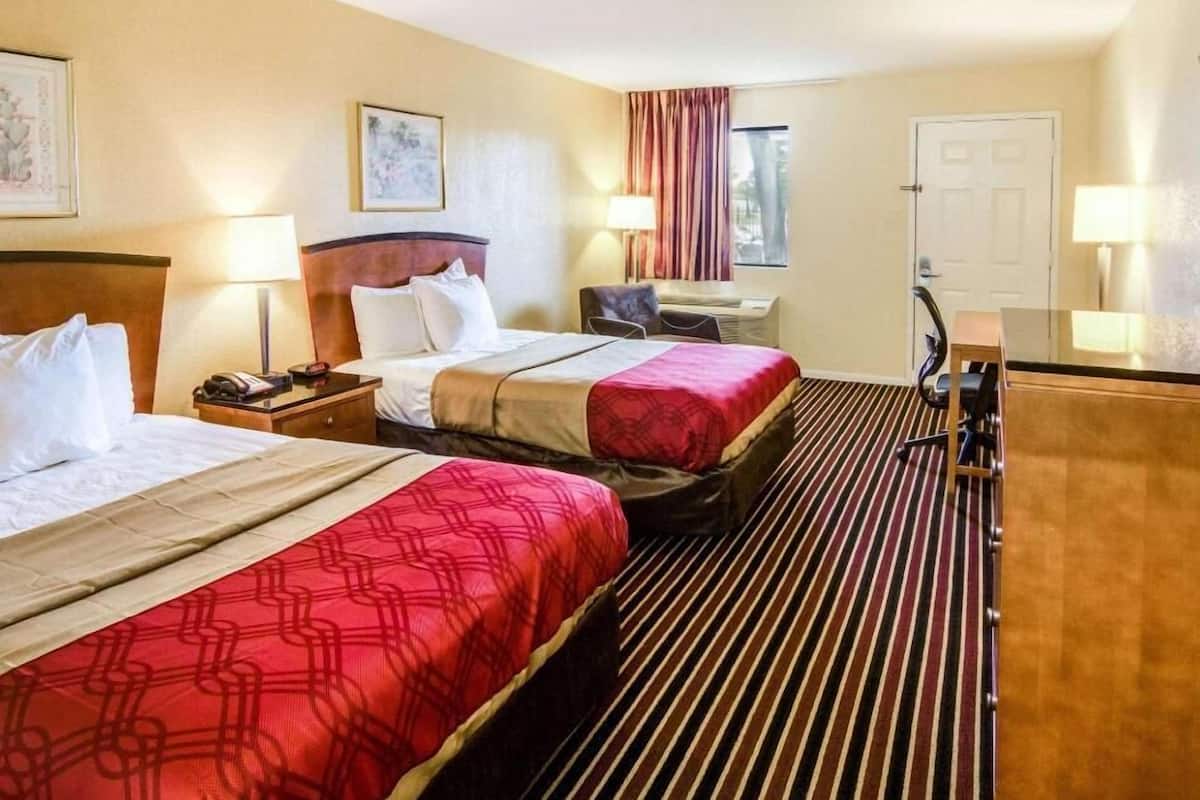 A hotel room with two beds, a desk, a chair, a lamp, and a window with curtains.