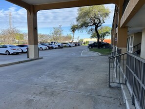 Un parking avec plusieurs voitures, l’entrée d’un immeuble et un arbre.