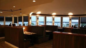 Ein Restaurant mit Holzwänden, Tischen und Sitzgelegenheiten, mit großen Fenstern und Blick auf das Wasser und die Skyline.