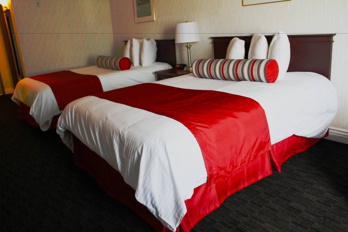 Ein Hotelzimmer mit zwei Betten, jeweils mit weißer Bettwäsche und roten Akzenten, einem Nachttisch mit Lampe und einem dunklen Teppichboden.