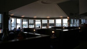 Ein modernes Interieur mit gewölbter Decke, großen Fenstern mit Blick auf die Stadt und Sitzbereichen mit Tischen und Stühlen.