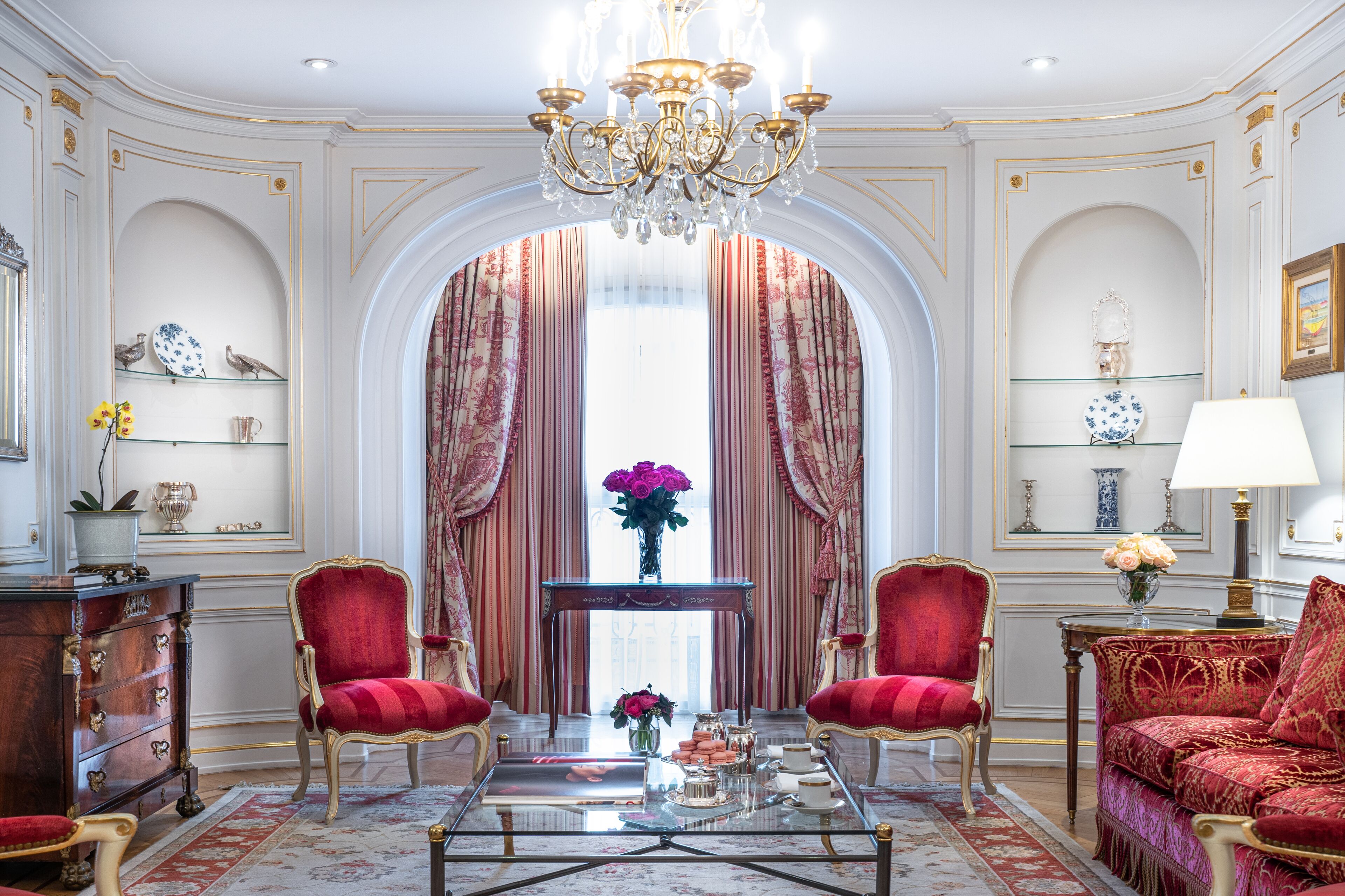 A luxurious room with a grand chandelier, red upholstered chairs, a coffee table, and a view through an arched doorway.