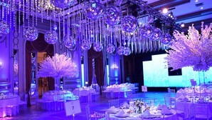 Un salón de banquetes con mesas redondas dispuestas para un evento formal, candelabros y un escenario con una pantalla grande.