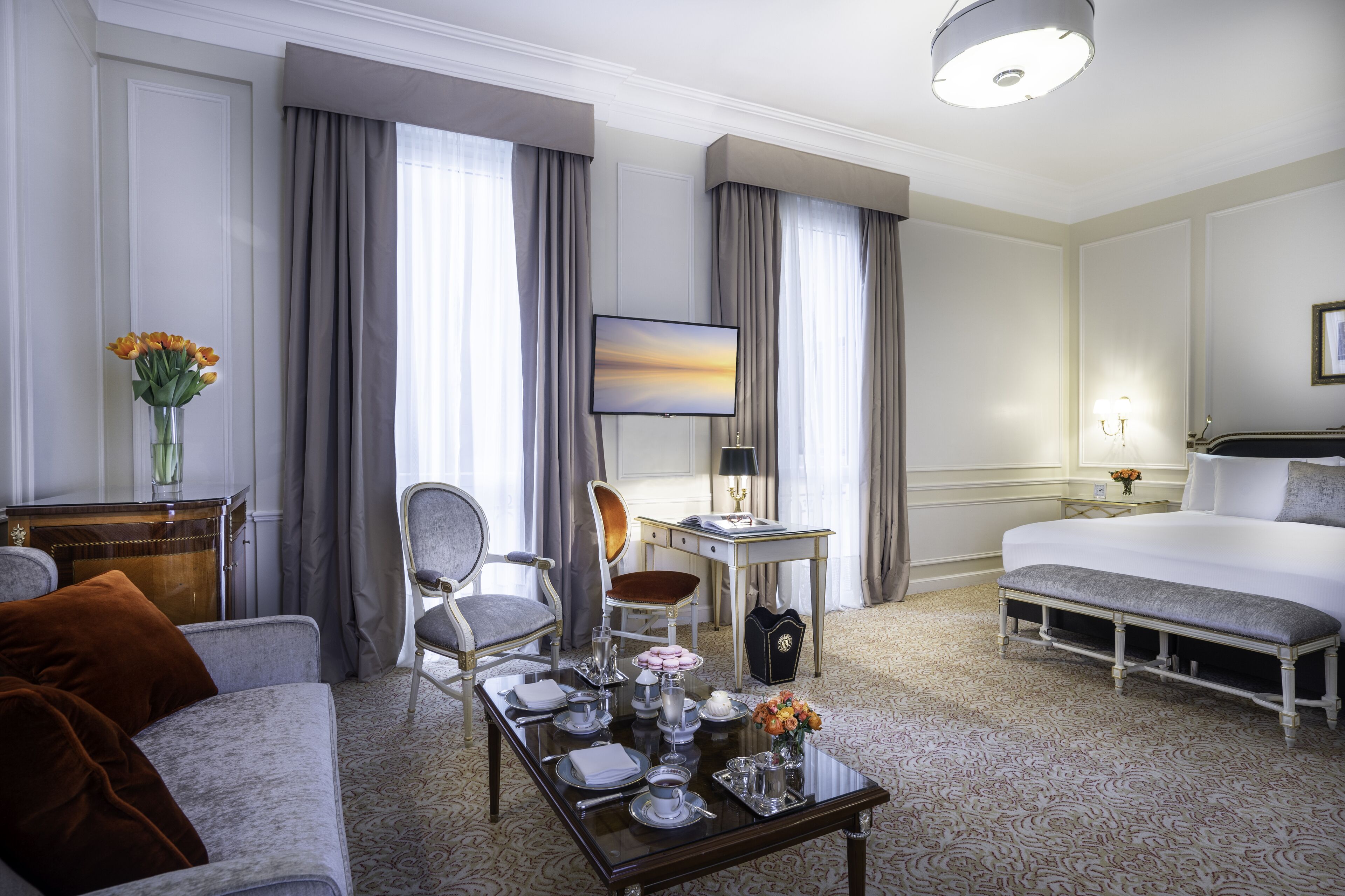 A hotel room with a bed, a sitting area with a sofa and armchairs, a dining table set for breakfast, and a television on a desk.