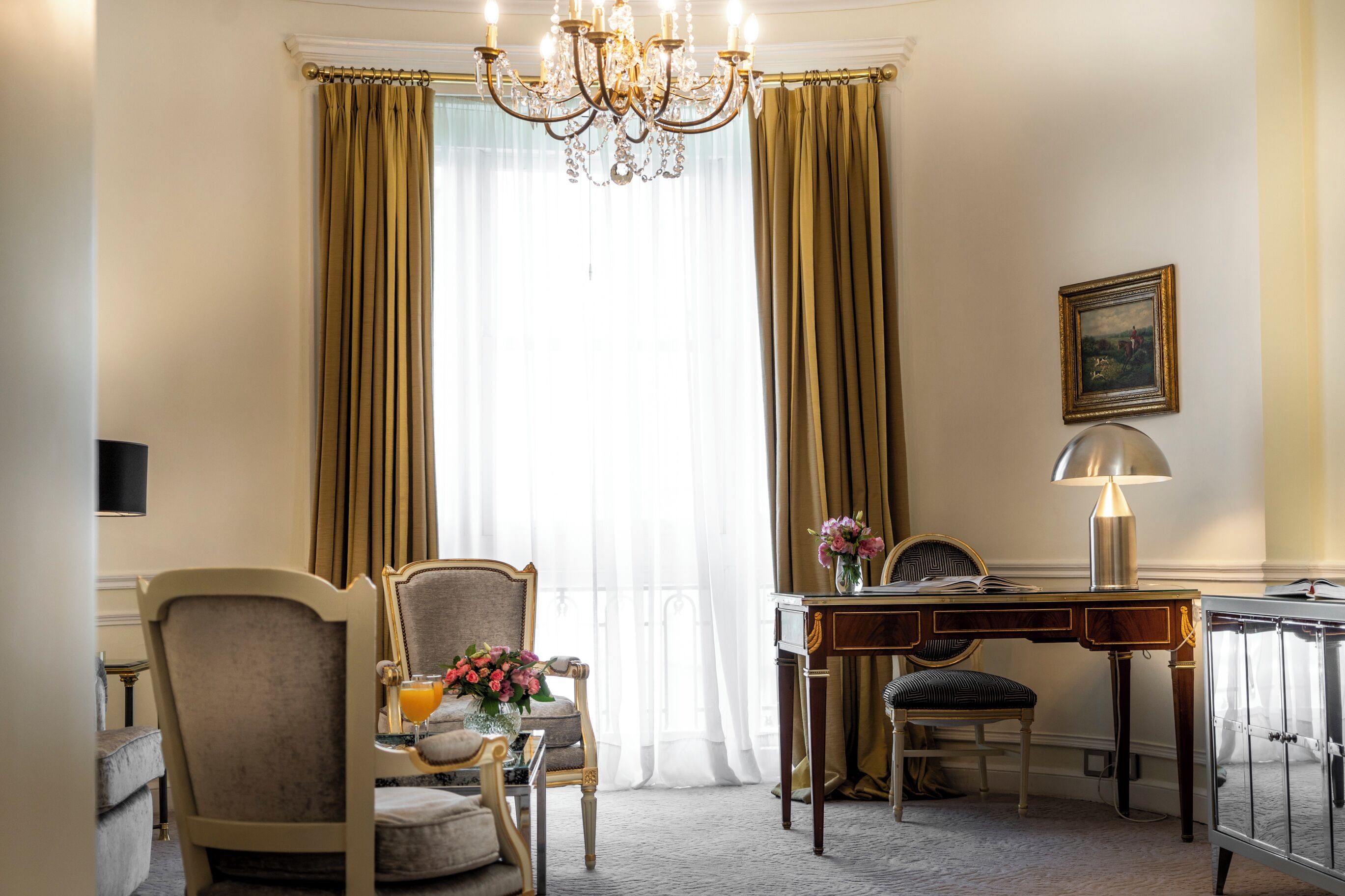 Ein Hotelzimmer mit großem Fenster, einer Kristallleuchtre, einem Schreibtisch mit Lampe, einem Sessel und einem kleinen Tisch mit Blumenvase.