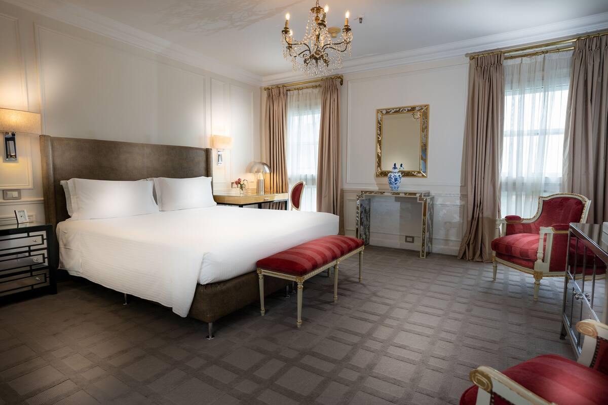Una habitación de hotel amplia con una cama grande, un banco, dos sillas, un tocador con espejo y una araña.
