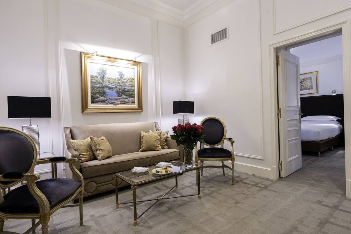 Una habitación de hotel con un sofá color beige, una mesa de centro de vidrio con un jarrón de rosas rojas, dos sillones, una cama con ropa blanca y un cuadro en la pared.