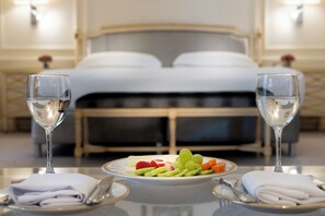 Una mesa de desayuno ordenada con una fuente de fruta, dos vasos de agua y servilletas y cubiertos doblados en una habitación de hotel con una cama al fondo.