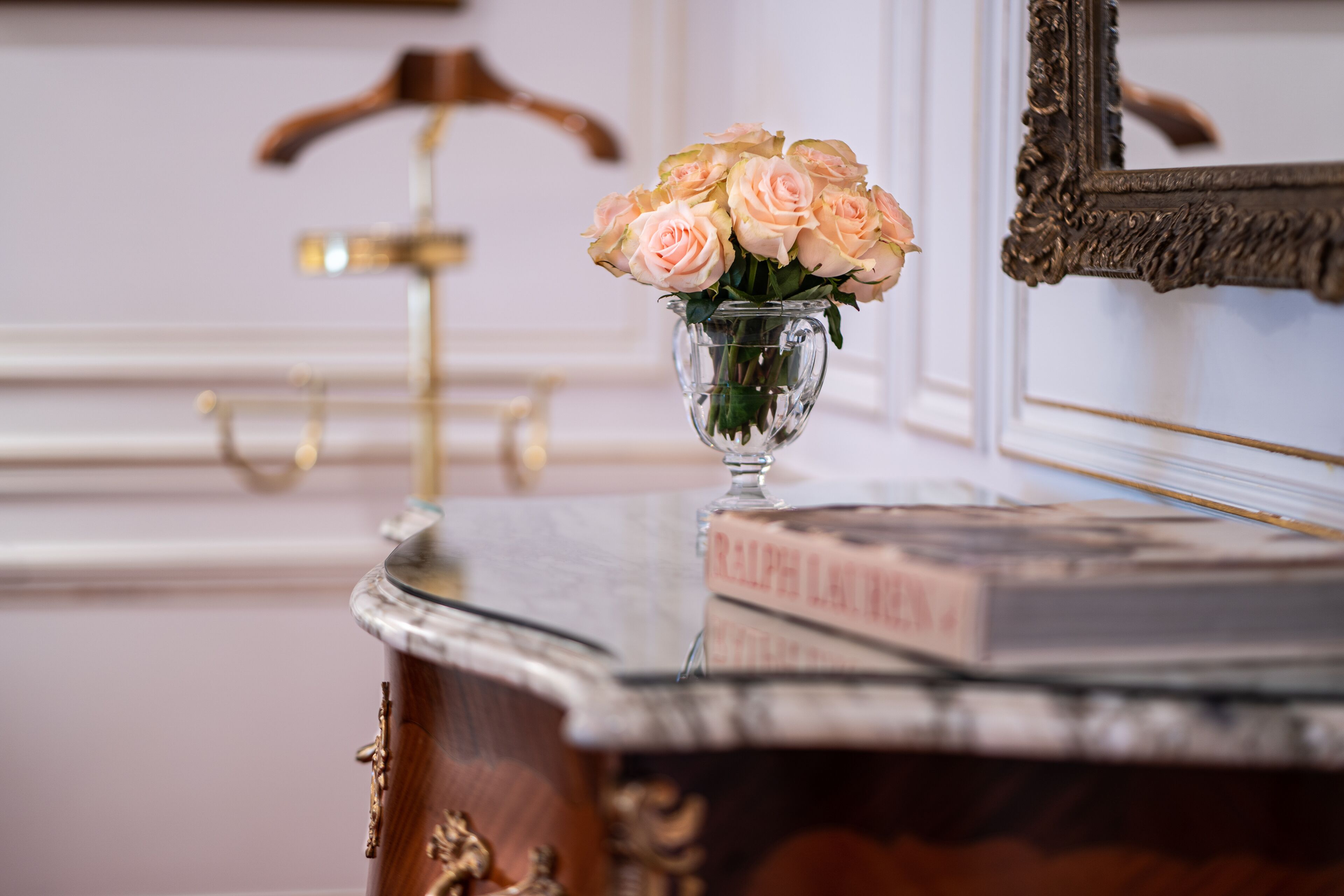 Una mesa de mármol con un florero de cristal con rosas rosadas, un espejo y un libro titulado "RALPH LAUREN".