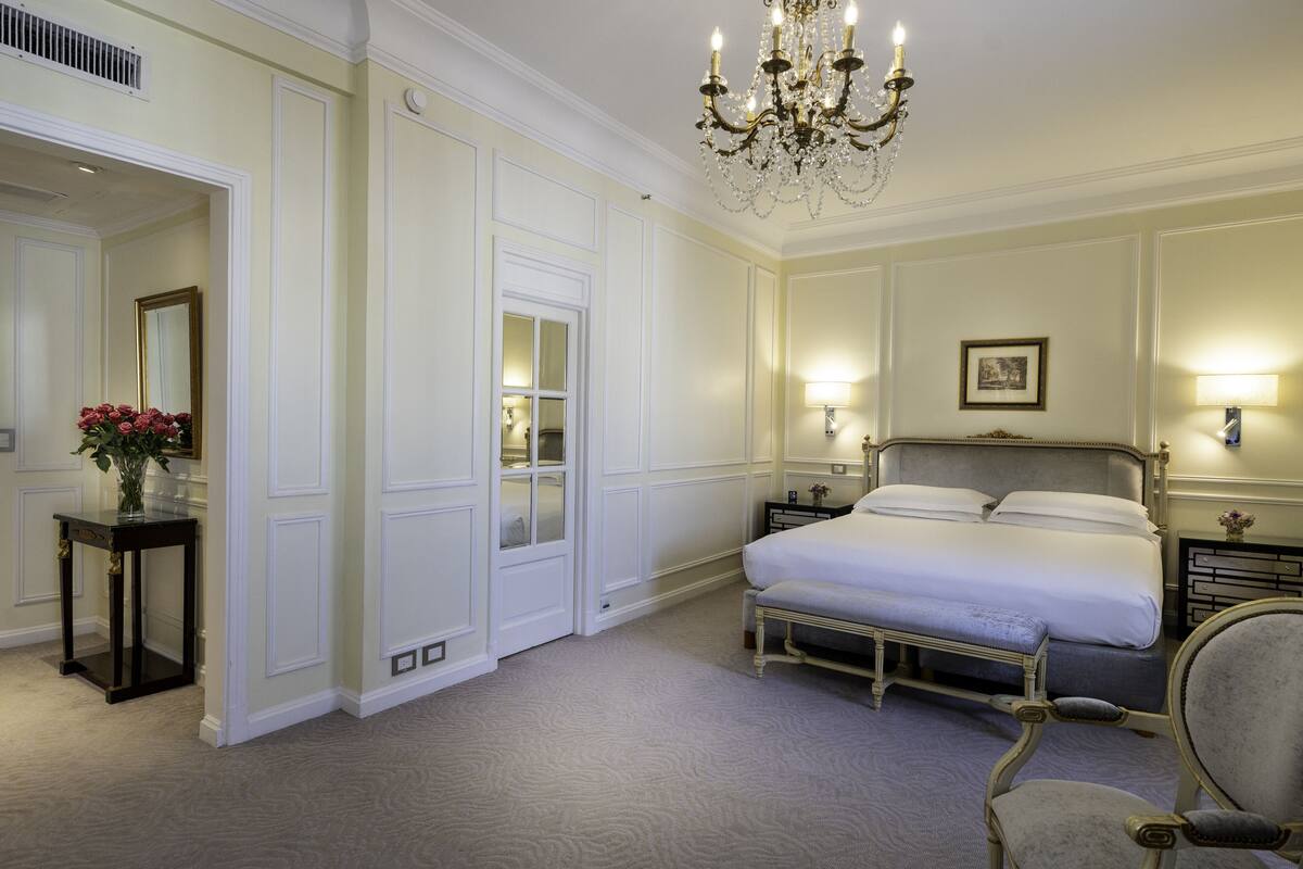 Una habitación de hotel con una cama grande, lámparas de noche, un candelabro, un espejo y una pequeña mesa con flores.