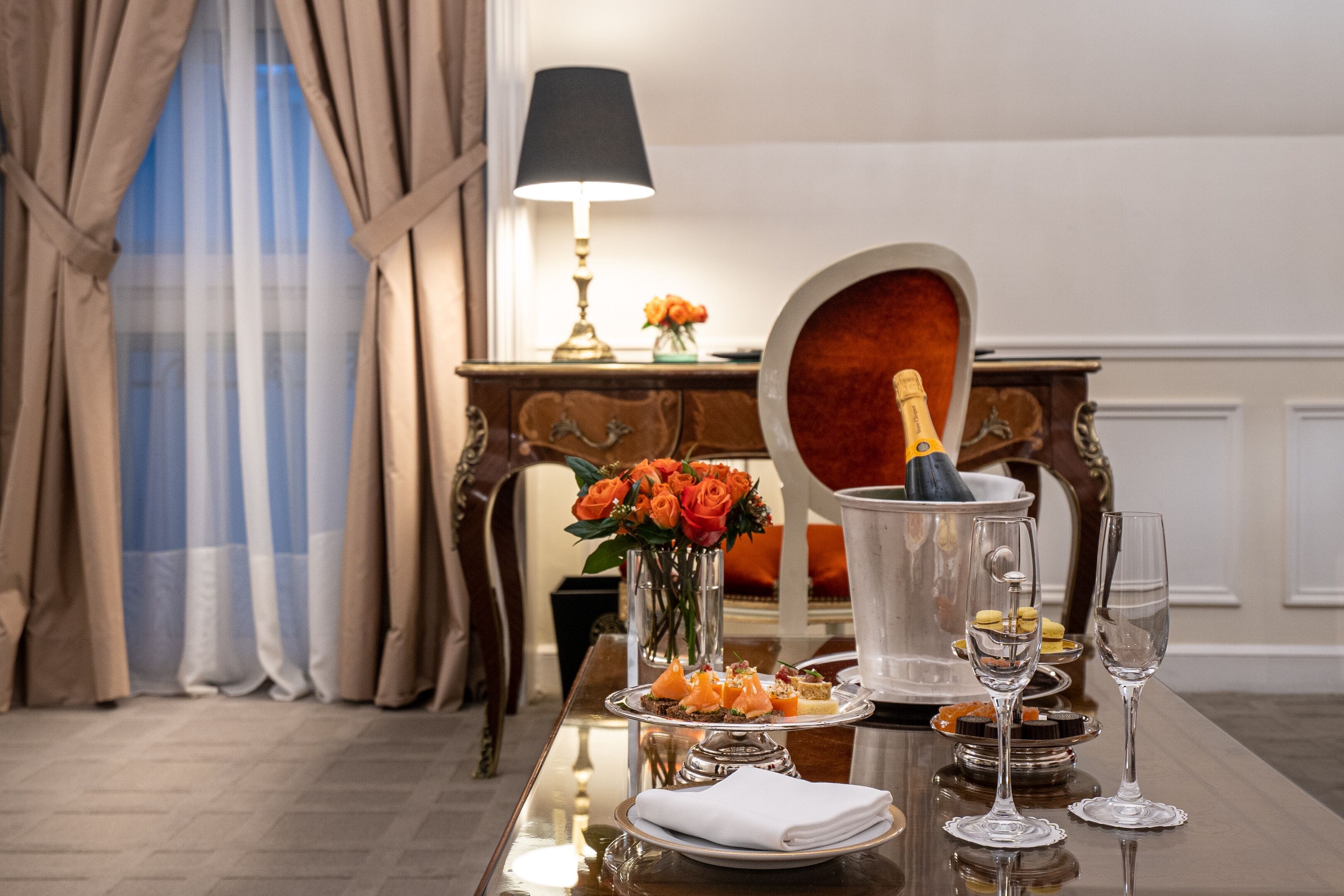 Una mesa de comedor con una botella de champán, un florero con flores naranjas y un plato de aperitivos.