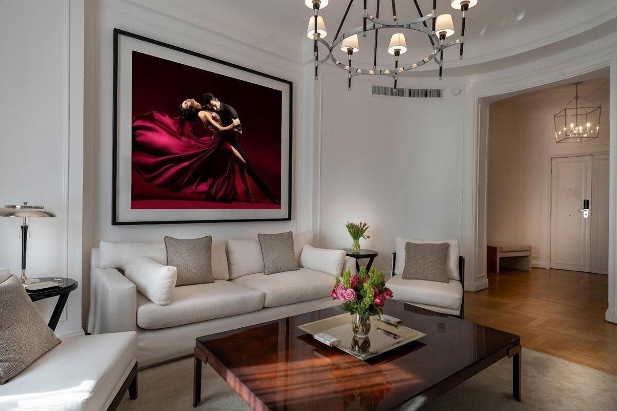 Una sala moderna con una obra de arte grande enmarcada, un conjunto de sofá blanco, una mesa de centro con un jarrón de flores y una araña.