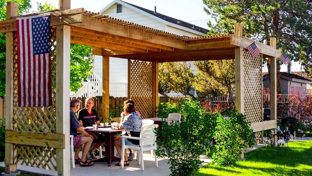 A wooden pergola with a table and chairs, surrounded by greenery and American flags.