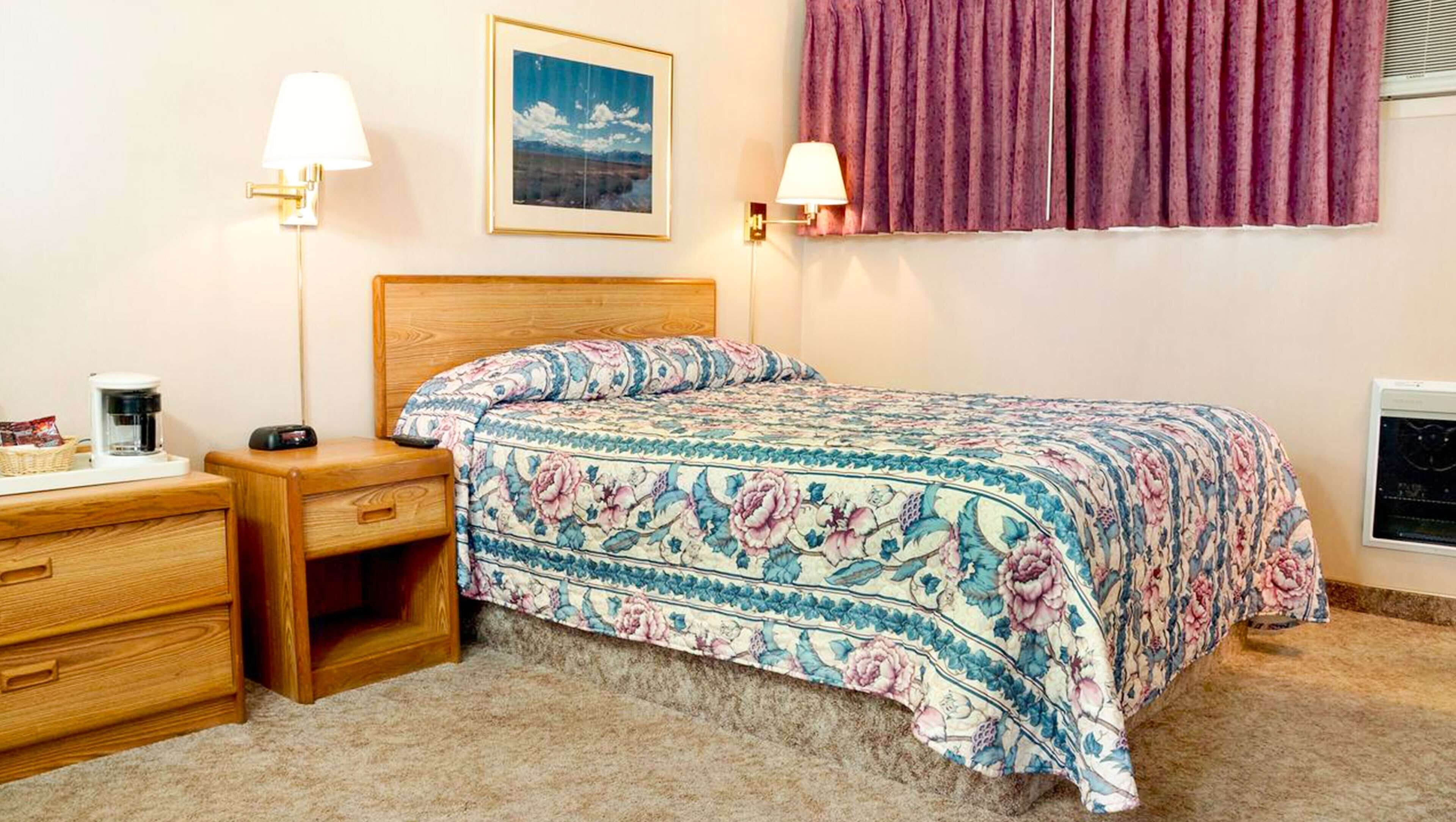 A bedroom with a wooden headboard, a floral bedspread, two bedside lamps, a coffee maker, and a framed picture on the wall.