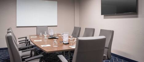 Una sala de conferencias con una gran mesa, seis sillas, una pantalla de proyector y un televisor de pantalla plana.