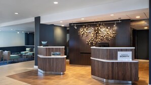 Un espace d’accueil moderne comprenant deux comptoirs de réception en bois, un coin salon avec des chaises et des tables, et un mur décoratif agrémenté de luminaires suspendus.