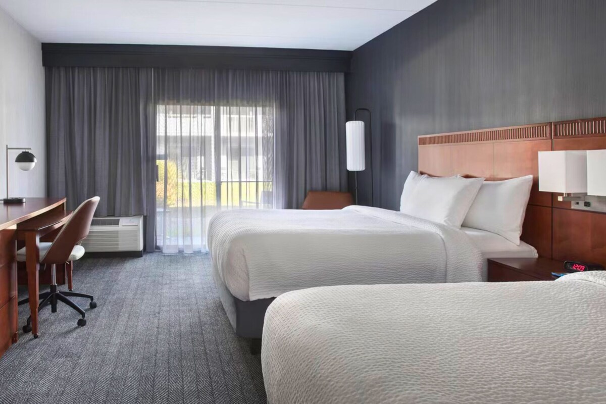 Habitación de hotel con escritorio, dos camas, una silla y un ventanal con cortinas.