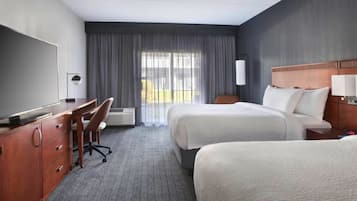 A hotel room with a desk, two beds, a chair, and a large window with curtains.