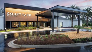Un hôtel Courtyard by Marriott doté d’un espace extérieur couvert avec des sièges, d’un jardin aménagé et d’une allée mouillée qui reflète le ciel.