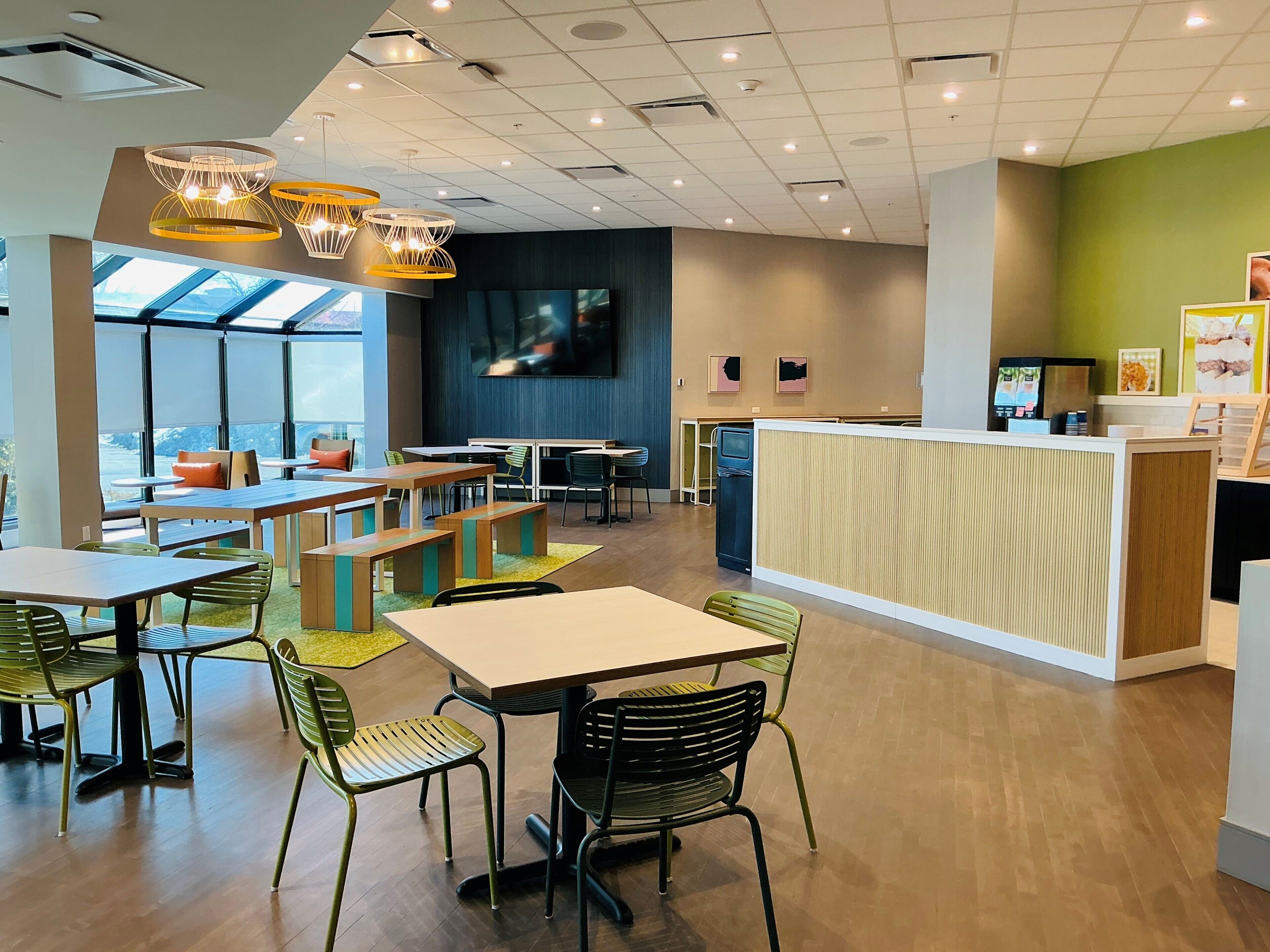 A modern cafe with wooden tables, green chairs, a counter, and a TV screen.