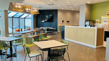 A modern cafe with wooden tables, green chairs, a counter, and a TV screen.