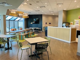 Una cafetería moderna con mesas de madera, sillas verdes, una barra y una pantalla de televisión.