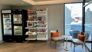 A modern interior with a refrigerator, shelves stocked with snacks, and a seating area by a window. - Spark by Hilton Montreal Brossard (Brossard)