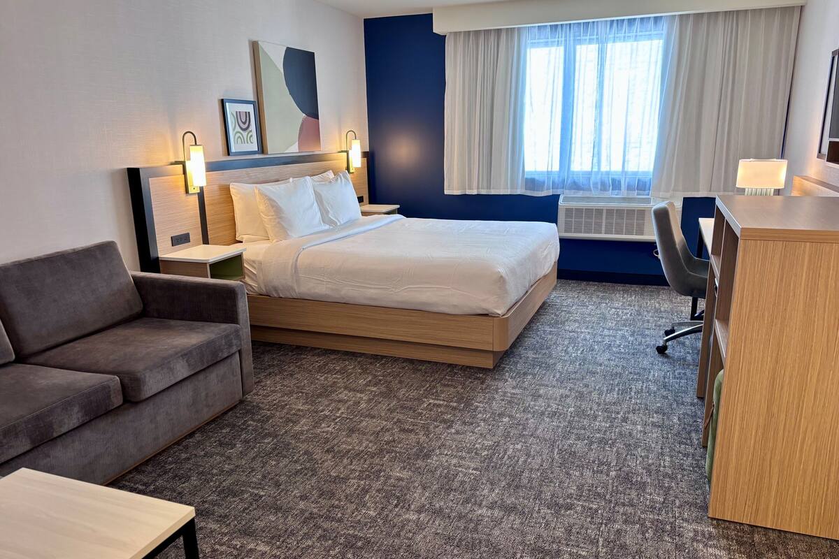A hotel room with a bed, a desk, a chair, a sofa, and a window with curtains.
