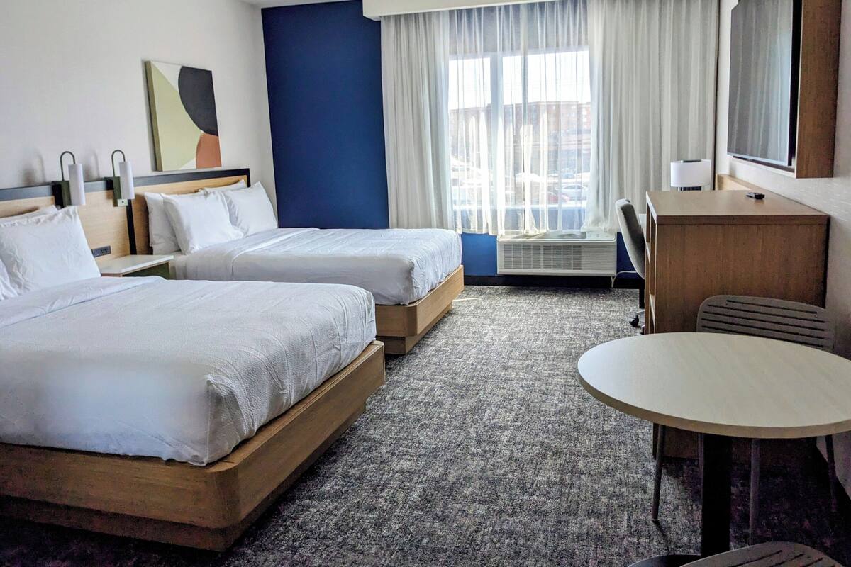 A hotel room with two beds, a desk, a chair, a TV, and a large window with curtains.