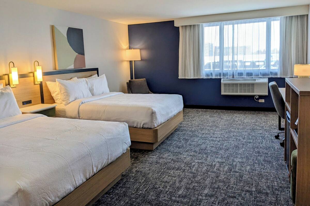 A hotel room with two beds, a desk, a chair, a lamp, and a window with curtains.