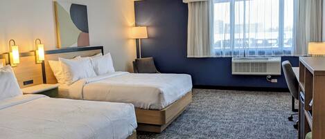 Habitación de hotel con dos camas, un escritorio, una silla, una lámpara y una ventana con cortinas.