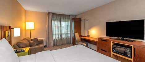 Una camera d'albergo con un letto, una scrivania con televisore a schermo piatto, un forno a microonde, una sedia, un comodino e una lampada.