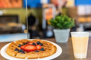 Un gofre cubierto de fresas y arándanos, un vaso de una bebida de color claro, y una planta en maceta al fondo.