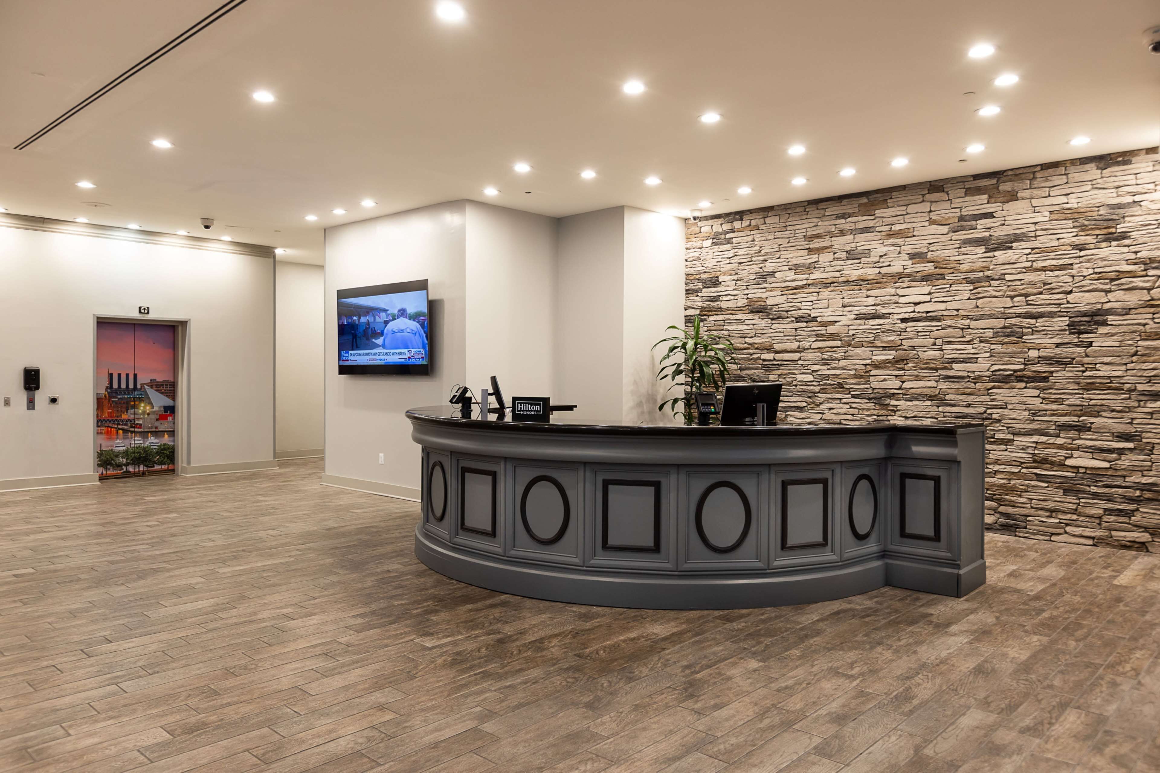 A hotel reception area with a curved grey reception desk, a stone wall, and a mounted television on the wall.