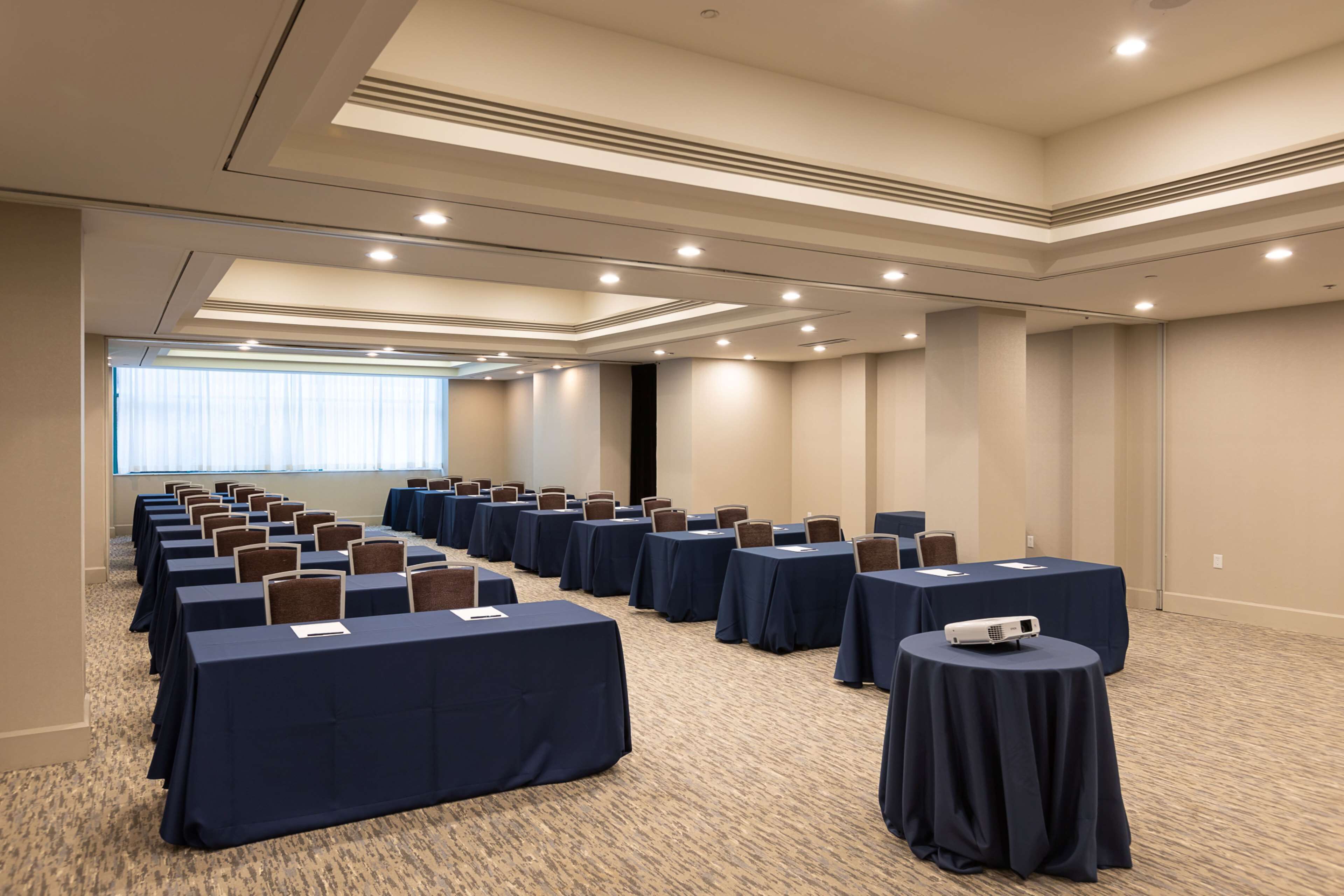 A conference room with rows of tables and chairs, a projector, and a large window with blinds.