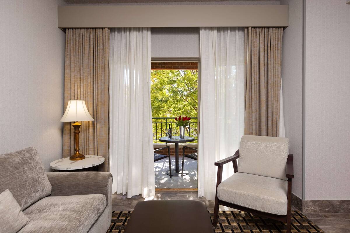 A hotel room with a sofa, armchair, lamp, and a view of an outdoor patio with a table and chairs.
